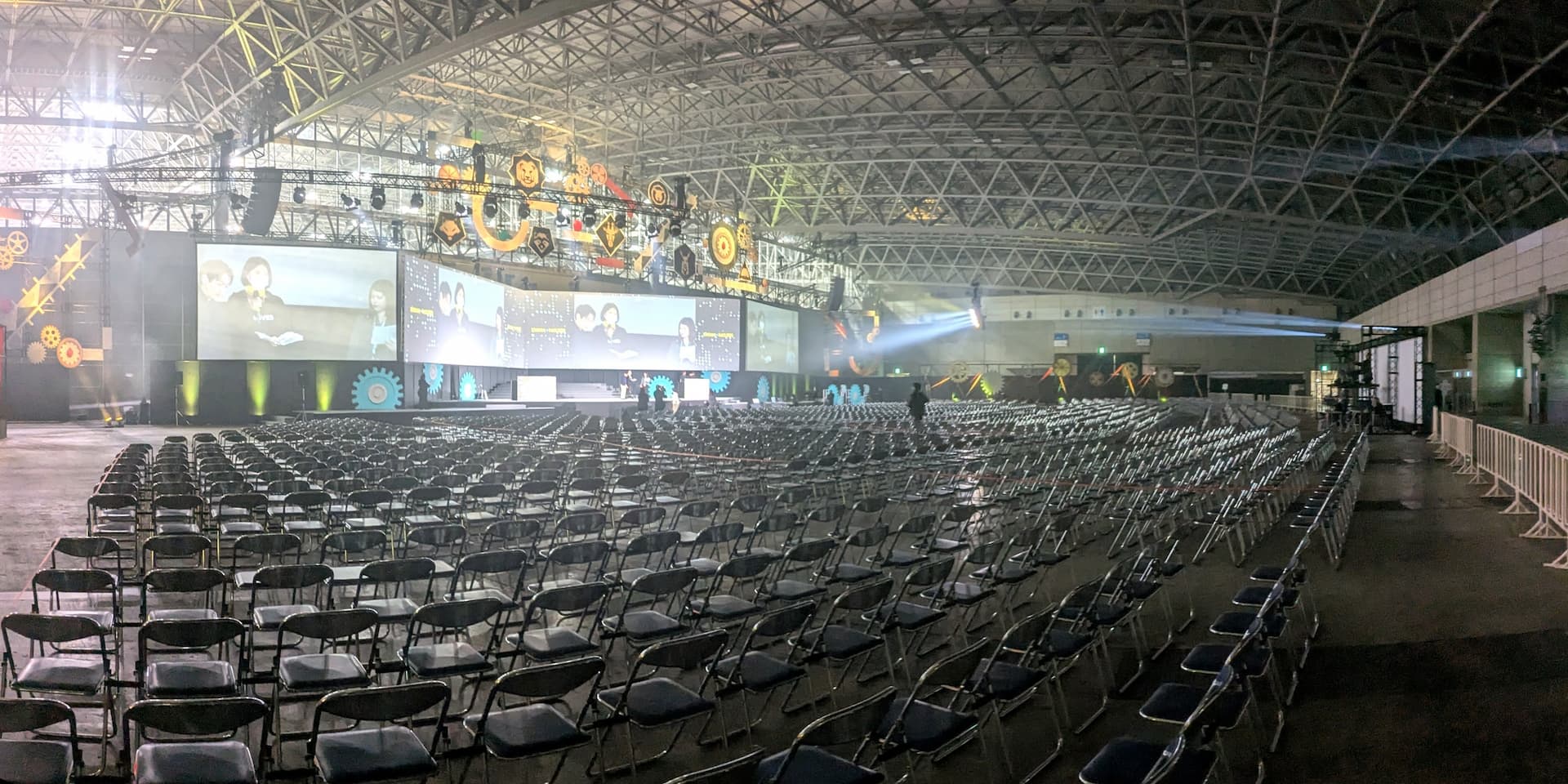 リハーサル中のステージを後ろから写した写真。屏風状に折れ曲がった巨大なスクリーンと、どこまでも続くパイプ椅子が写っている。